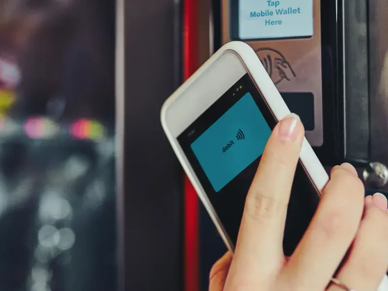 A hand holding a phone with a debit card on screen up to a vending machine reader marked Tap Mobile Wallet Here
